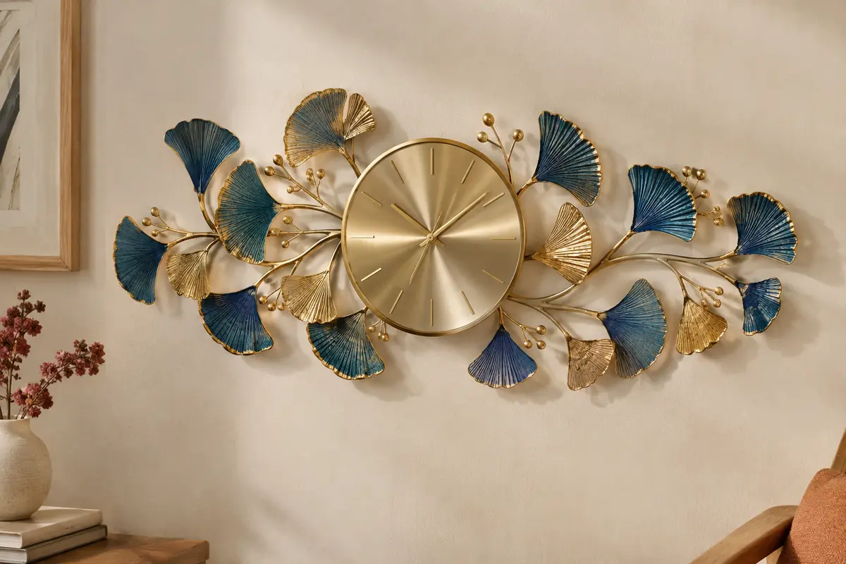Horloge murale en métal avec branches dorées et feuilles de ginkgo bleues et dorées sur un mur crème.