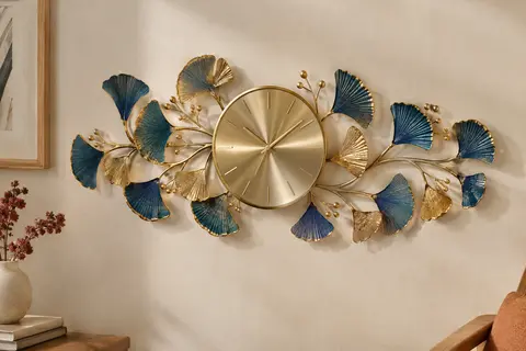 Horloge murale en métal avec branches dorées et feuilles de ginkgo bleues et dorées sur un mur crème.