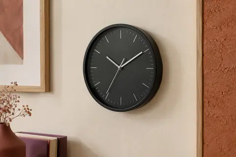 Horloge murale ronde noire avec aiguilles argentées, présentée sur un mur crème près d’un cadre en bois clair.